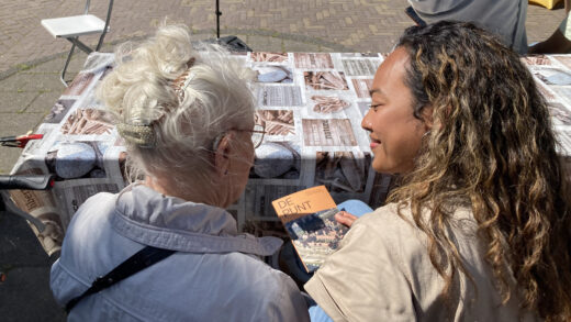 Twee vrouwen zittend aan een tafel, van achteren gefotografeerd. Buurtbewoonster Gré krijgt het boekje over De Punt van een communicatiemedewerker van Lieven de Key.